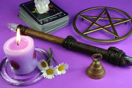 Magic tools: a pink candle, wooden and crystal wand, pentagram, tarot cards, crystal and a bell on a purple background.